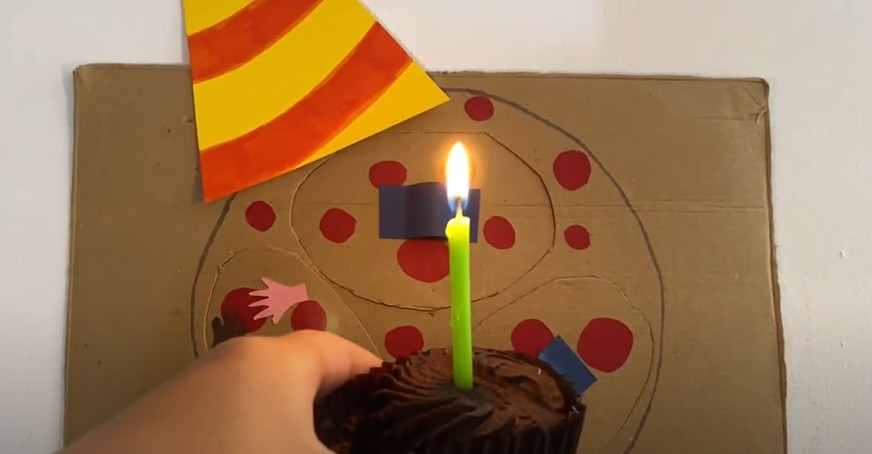 Screenshot from video of cardboard cut-out of person with chickenpox wearing a party hat and holding a cupcake with one lit candle 