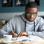 Student with glasses writing in a book with laptop and mug of coffee infront