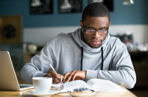 Student with glasses writing in a book with laptop and mug of coffee infront