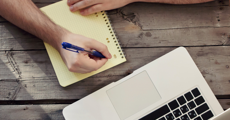 person writing on a yellow page in front of laptop