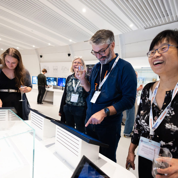 Main image Group of people exploring interactive displays in a modern, well-lit exhibition space with white walls and ceiling panels. Secondary image is group of audience members in a lecture hall clapping.