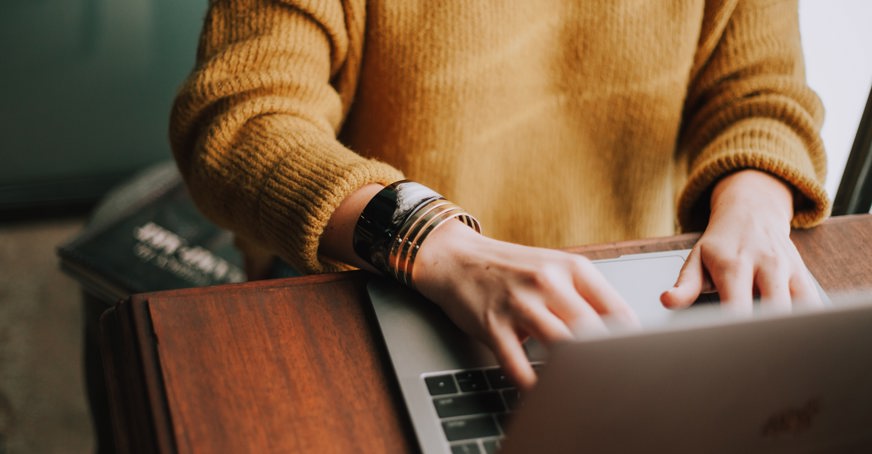 Image of person in yellow jumper typing on laptop at wooden desk