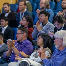 Main image of attendees asking questions from their seats with microphone. Secondary image is of group of attendees in a lecture hall clapping.