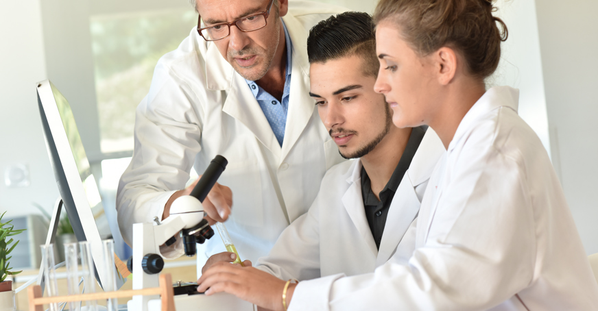 Teacher in a lab coat helping students in lab coats to use a microscope