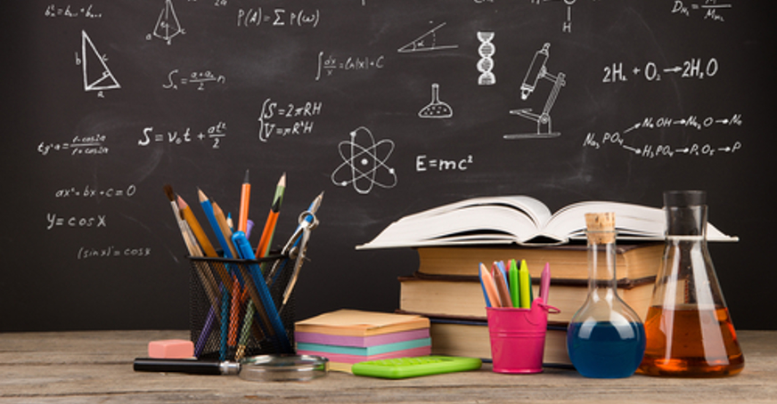 Blackboard with scientific writing behind a table full of books, pencils and scientific flasks