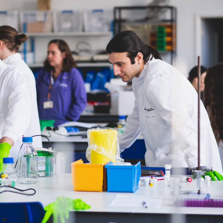 Main image of scientists in white lab coats and green gloves conducting experiments with lab equipment on a cluttered table in a busy laboratory. Secondary image is of group of attendees in a room with poster boards on display.