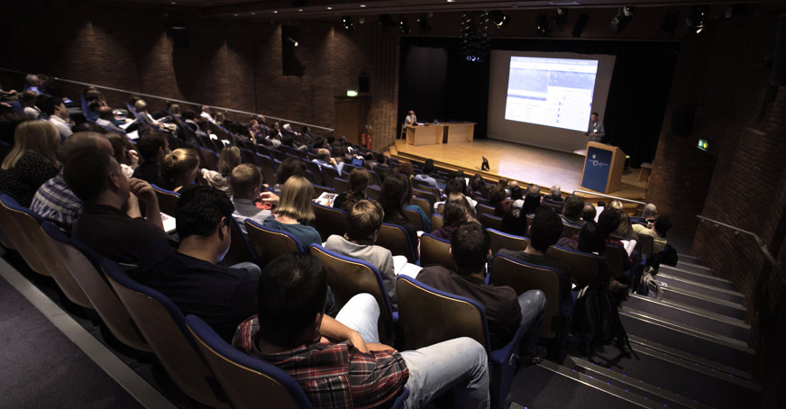 View from the back of lecture theater with students in the seats and a lecturer on stage in front of  a screen