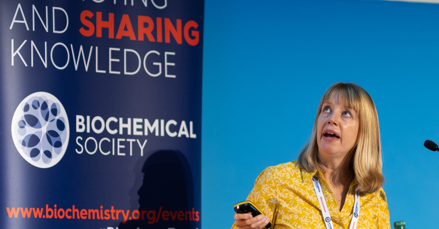 Speaker holding a laser pointer at a screen standnig in front of Biochemical Soceity banner