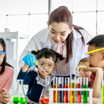 Female teacher in a lab coat showing three young children how to conduct a science experiment with testtubes